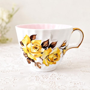 May include: A white porcelain teacup featuring a fluted design and a soft pink interior. The exterior is decorated with two large yellow roses with brown leaves and stems. The cup includes a gold-trimmed handle and a gold-painted rim.