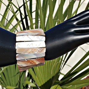 Puede incluir: Un brazalete ancho hecho de piezas rectangulares e iridiscentes de concha en tonos blancos, marrones y beige. El brazalete se muestra en una mano de maniquí negra sobre un fondo de hojas de palmera verdes.