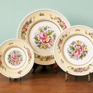May include: Three antique decorative plates with floral designs. The plates have a cream-colored background with a central bouquet of pink, yellow, and blue flowers. The rims are adorned with a beaded edge and additional floral accents. The plates are displayed on small stands.