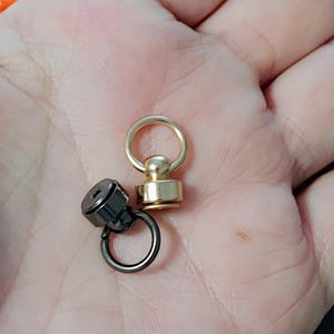 May include: Two swivel snap hooks, one gold and one black, with a round ring on each. The hooks are on a hand.