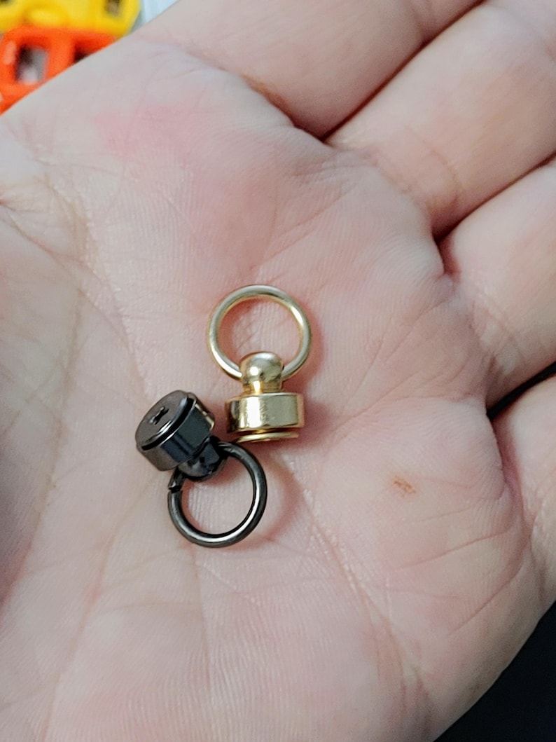 May include: Two swivel snap hooks, one gold and one black, with a round ring on each. The hooks are on a hand.