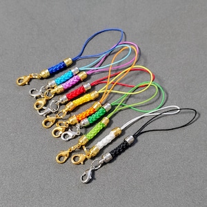 May include: A set of ten colorful braided phone charms with gold and silver clasps. Each charm is a different color, including blue, pink, red, orange, yellow, green, white, and black.