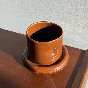 May include: A brown ceramic cup and saucer set. The cup is cylindrical with a smooth, glossy finish and sits on a matching saucer. The set is a warm, earthy brown color, perfect for serving coffee or tea.