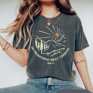 May include: Dark gray t-shirt with a mountain and sun graphic. The design includes trees, mountains, and the text "IN PRINCIPIO ERAT VERBUM" and "JOHN 1:1". The shirt is a relaxed fit and has a vintage look.