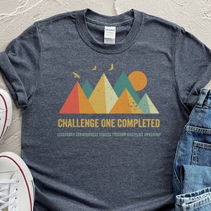 May include: A dark heather grey t-shirt with a retro mountain graphic and the text "Challenge One Completed: Leadership, Confidence, Choice, Freedom, Discipline, Ownership".