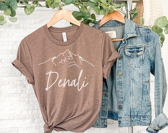 May include: A brown t-shirt with a white outline of a mountain range and the word "Denali" in white script. The t-shirt is hanging on a rack with a denim jacket.