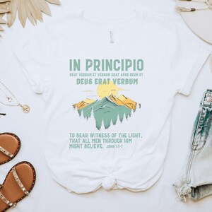 May include: White t-shirt featuring the text "IN PRINCIPIO" and a mountain landscape design in green and yellow. The shirt also includes a quote from John 1:1-7. A casual, everyday wear tee.