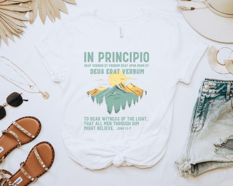 May include: White t-shirt featuring the text "IN PRINCIPIO" and a mountain landscape design in green and yellow. The shirt also includes a quote from John 1:1-7. A casual, everyday wear tee.