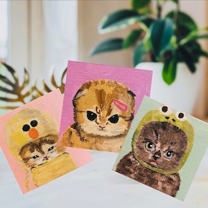 May include: Three hand-painted canvases featuring adorable kittens wearing different hats. The first canvas shows a brown tabby kitten wearing a pink headband. The second canvas shows a white kitten wearing a yellow chick hat. The third canvas shows a black kitten wearing a green frog hat.