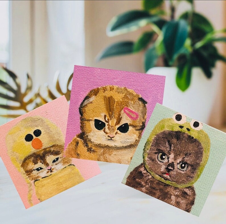 May include: Three hand-painted canvases featuring adorable kittens wearing different hats. The first canvas shows a brown tabby kitten wearing a pink headband. The second canvas shows a white kitten wearing a yellow chick hat. The third canvas shows a black kitten wearing a green frog hat.