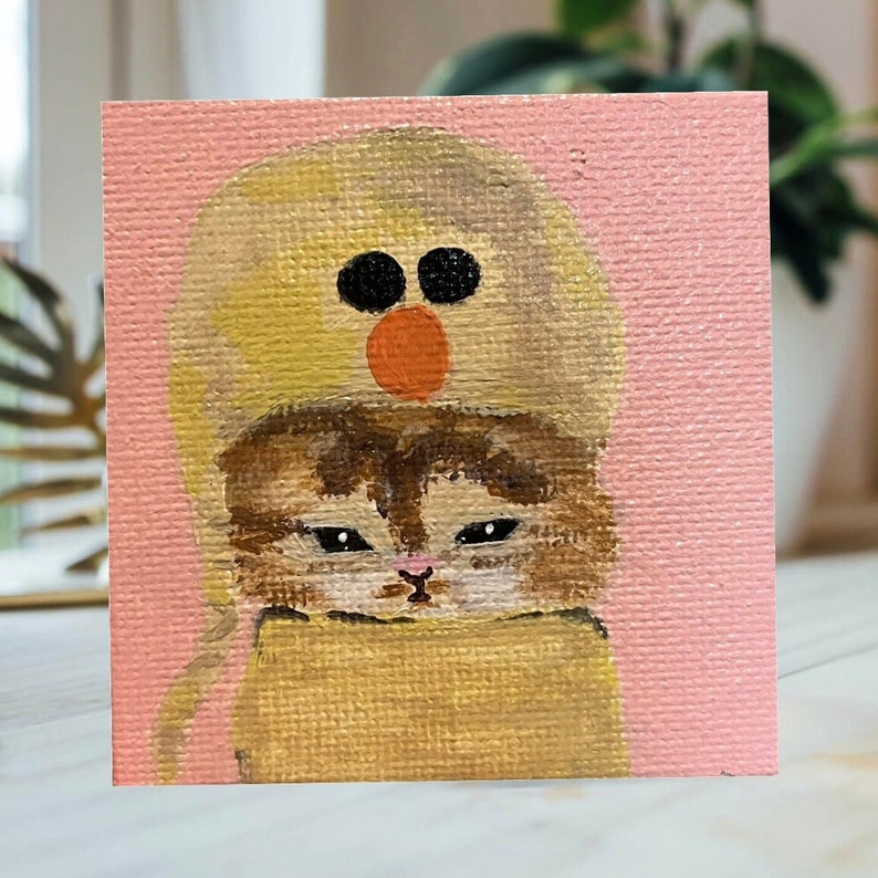 May include: A painting of a brown tabby cat wearing a yellow hat with a red and black face on it. The cat is looking down and the background is pink.