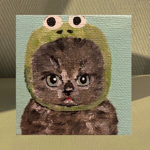 May include: A painting of a grey cat wearing a green frog hat with large, white eyes. The cat has a serious expression and is looking directly at the viewer.