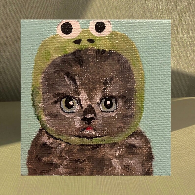 May include: A painting of a grey cat wearing a green frog hat with large, white eyes. The cat has a serious expression and is looking directly at the viewer.