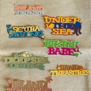 May include: A collection of colorful, layered wooden signs with beach-themed phrases. Phrases include "Island Drinks," "Scuba Diving," "Under the Sea," "Bikini Babe," "Deep Sea Diving," "Sand Treasures," and "Island Retreat."