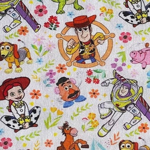 May include: A white fabric with a repeating pattern of Toy Story characters, including Woody, Buzz Lightyear, Jessie, and Rex. The characters are surrounded by colourful flowers and leaves.