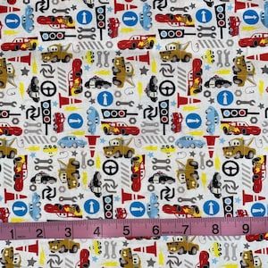 May include: White fabric with a repeating pattern of cartoon cars, traffic lights, wrenches, and steering wheels. The cars are in various colors, including red, yellow, and blue. A pink measuring tape shows 9 inches.