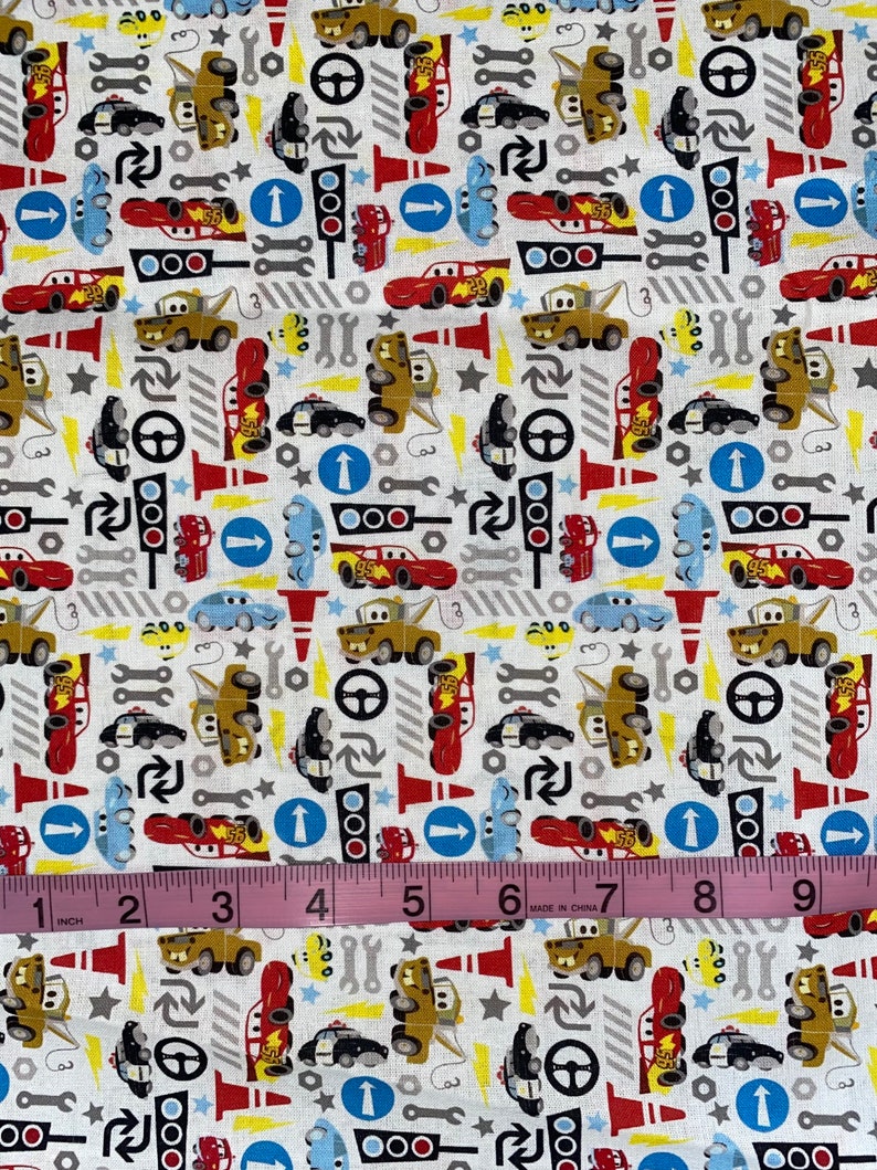 May include: White fabric with a repeating pattern of cartoon cars, traffic lights, wrenches, and steering wheels. The cars are in various colors, including red, yellow, and blue. A pink measuring tape shows 9 inches.