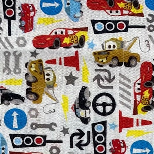 May include: White fabric with a repeating pattern of cartoon cars, traffic lights, wrenches, and directional signs. The cars are red, blue, and yellow. The traffic lights are red, yellow, and green. The directional signs are blue with white arrows.