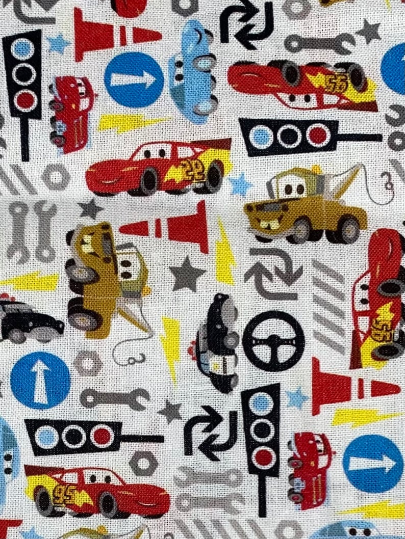 May include: White fabric with a repeating pattern of cartoon cars, traffic lights, wrenches, and directional signs. The cars are red, blue, and yellow. The traffic lights are red, yellow, and green. The directional signs are blue with white arrows.