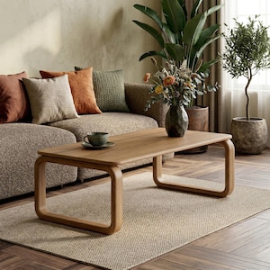 Peut inclure: Une table basse rectangulaire en chêne clair avec des coins et des pieds arrondis. La table est posée sur un tapis beige devant un canapé marron avec des coussins décoratifs. Un vase de fleurs et une tasse à thé sont sur la table.
