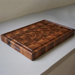 Puede incluir: Una tabla de cortar rectangular de madera marrón oscuro. La tabla presenta una ranura para jugos alrededor del perímetro y agarres para los dedos en los lados. El patrón de la veta de la madera es visible en toda la tabla.