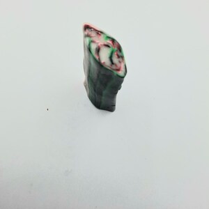 May include: A small, abstract sculpture with a layered design. The top edge is pink, transitioning to white, with green and red swirls. The sides are dark grey, resembling a stone or mineral.