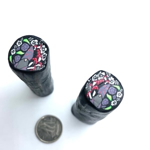 May include: Two black polymer clay canes with a colorful floral and geometric design. The design features white, red, green, purple, and gray colors.