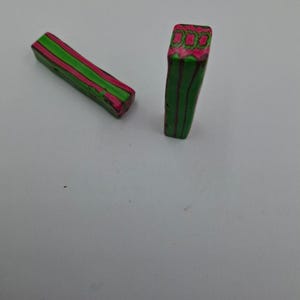 May include: Two rectangular polymer clay beads. One is lying down, the other is standing up. Both are green with pink and red stripes. The top of the standing bead has a pink and red pattern.
