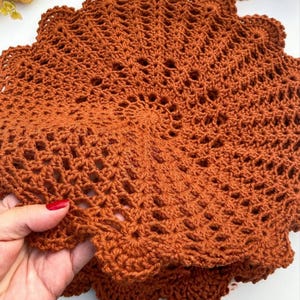 Puede incluir: Primer plano de un tapete de ganchillo hecho a mano, de color óxido, con un borde festoneado. El intrincado diseño calado es visible en toda la pieza circular. El tapete está sostenido por una mano con esmalte de uñas rojo.
