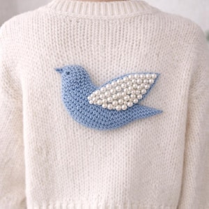 Op de afbeelding: Crèmekleurige gebreide trui met een lichtblauwe vogelapplicatie. De vogel heeft een gevleugelde vleugel, die een decoratief element toevoegt aan het gezellige kledingstuk met lange mouwen. De trui is een klassieke stijl, geschikt voor vrijetijdskleding.