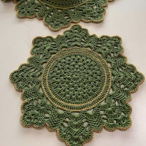 Może przedstawiać: Dwa zielone szydełkowe podkładki pod talerze z falistym brzegiem i złotym wykończeniem. Środek każdej podkładki ma okrągły wzór, również ze złotym wykończeniem. Podkładki są ręcznie robione i mają szczegółowy wzór.