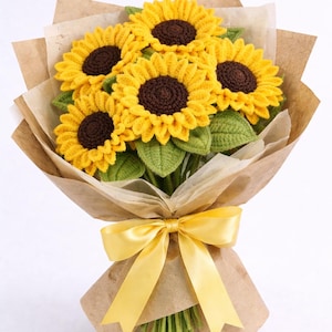 Puede incluir: Un ramo de girasoles de ganchillo con pétalos amarillos y centros marrones, envuelto en papel marrón y atado con una cinta amarilla. El ramo incluye hojas y tallos verdes, creando un arreglo floral vibrante y artesanal.