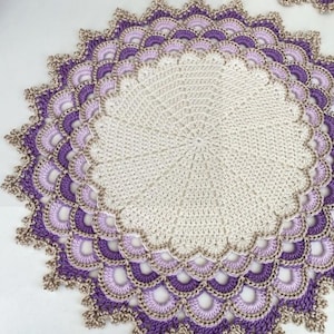Può includere: Un centrino rotondo all'uncinetto fatto a mano nei toni del crema, lavanda e viola. Il centrino presenta un bordo smerlato con un design traforato dettagliato. Il bordo esterno è rifinito con un filo marrone a contrasto.