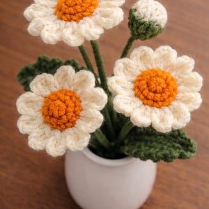 Peut inclure: Un arrangement de marguerites au crochet fait à la main dans un pot en céramique blanc. Les marguerites ont des pétales blancs et des centres orange. Des feuilles et des tiges vertes complètent le design floral. L'arrangement comprend trois fleurs ouvertes et un bouton.