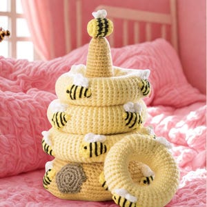 May include: A handmade yellow crochet stacking toy shaped like a beehive, with a small bee on top. The toy has several yellow rings with black and yellow bees attached. The base of the beehive has a brown honeycomb design.