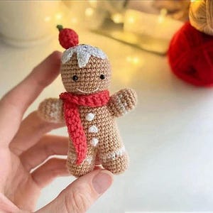 May include: A crocheted gingerbread man ornament with a red scarf and a berry on top. The gingerbread man has white icing details and black eyes. The ornament is held in a hand.