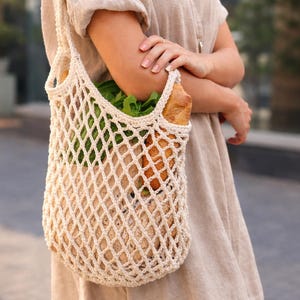 May include: A cream-colored, crocheted net shopping bag filled with fresh produce and a loaf of bread. The bag is held by a person wearing a beige dress. The bag's open weave design allows the contents to be visible.