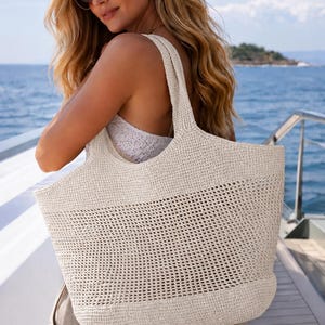 May include: A large, off-white tote bag crafted from woven material. The bag features a solid base, a wide middle section with an open-knit mesh pattern, and two sturdy shoulder straps. The design is simple, functional, and neutral in colour.