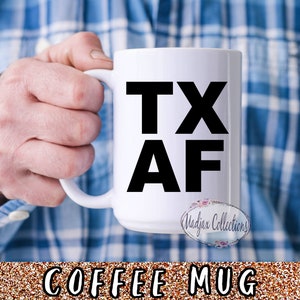 Könnte beinhalten: Eine weiße Kaffeetasse mit schwarzem Text, der "TX AF" lautet. Die Tasse wird von einer Hand gehalten, die ein blaues kariertes Hemd trägt.