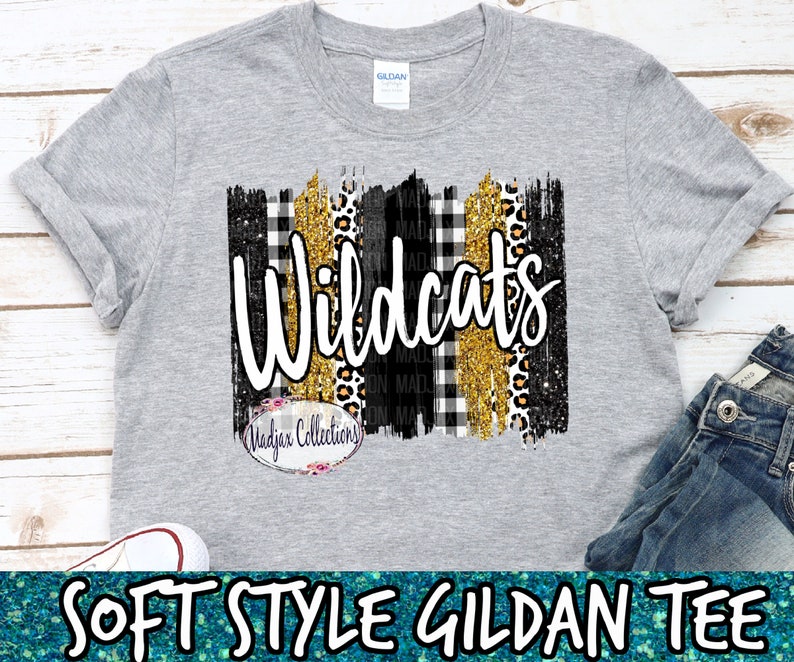 Op de afbeelding: Grijs T-shirt met een zwart, wit, goud en luipaardprint ontwerp. Het ontwerp bevat het woord "Wildcats" in witte tekst. Het shirt is een zacht Gildan T-shirt.