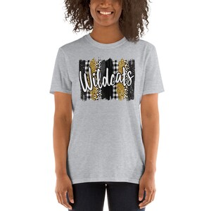 Op de afbeelding: Grijs T-shirt met korte mouwen met een zwart, wit en gouden grafisch ontwerp. De grafiek toont het woord "Wildcats" in witte letters met zwart-gouden ruitjespatronen, luipaardprint en gouden glitter.