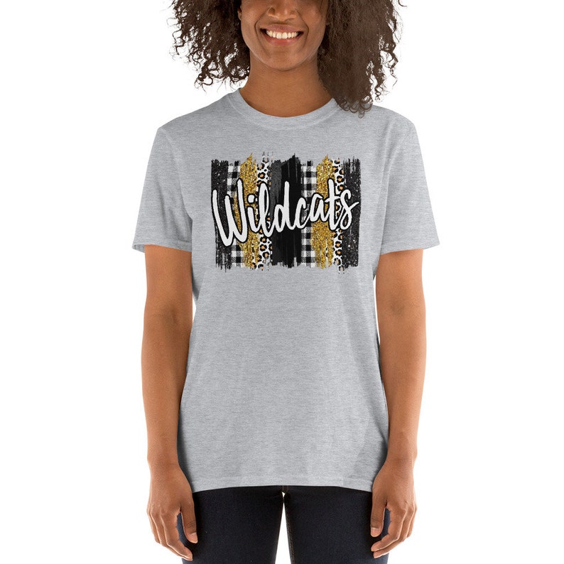Op de afbeelding: Grijs T-shirt met korte mouwen met een zwart, wit en gouden grafisch ontwerp. De grafiek toont het woord "Wildcats" in witte letters met zwart-gouden ruitjespatronen, luipaardprint en gouden glitter.