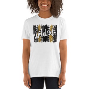 Op de afbeelding: Wit T-shirt met een zwart-wit geruit patroon en gouden glitteraccenten. Het woord "Wildcats" staat in zwarte schrift op het shirt.