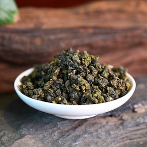 May include: A white ceramic bowl filled with loose green tea leaves. The tea leaves are small and tightly rolled, with a dark green color.