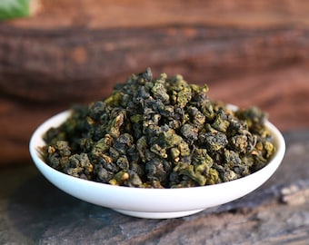May include: A small white ceramic bowl filled with a mound of tightly rolled, dark green oolong tea leaves. The leaves exhibit a textured, curled appearance with varying shades of deep forest green and hints of lighter olive green throughout.