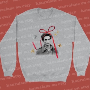 Puede incluir: Una sudadera gris claro con un retrato en blanco y negro, una cinta roja y elementos decorativos. El diseño incluye una concha, estrellas y una cruz. Esta sudadera informal es una prenda cómoda.