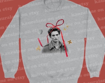 May include: A light gray sweatshirt featuring a black and white image of a person with a red ribbon and decorative elements. The design includes a seashell, stars, and a cross. The sweatshirt is a casual garment.