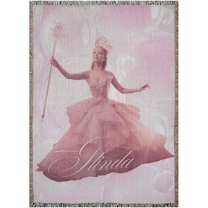 Peut inclure: Couverture tissée rose avec une illustration d'une femme en robe rose, tenant une baguette étoilée. Le nom "Glinda" est écrit en cursive. Le fond présente un motif doux en forme de bulles. Bords frangés.