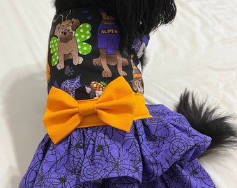 May include: A Halloween-themed dog dress featuring a black bodice with cartoon dog graphics and an orange bow. The skirt is purple with a black spiderweb pattern. The dress is designed for a small dog.