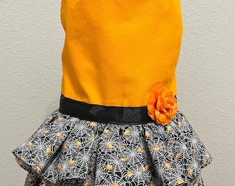 May include: A Halloween-themed pet dress. The top is bright orange, with a black ribbon belt and an orange flower. The skirt is black with a white spiderweb pattern and orange spiders. The dress has a halter top design.